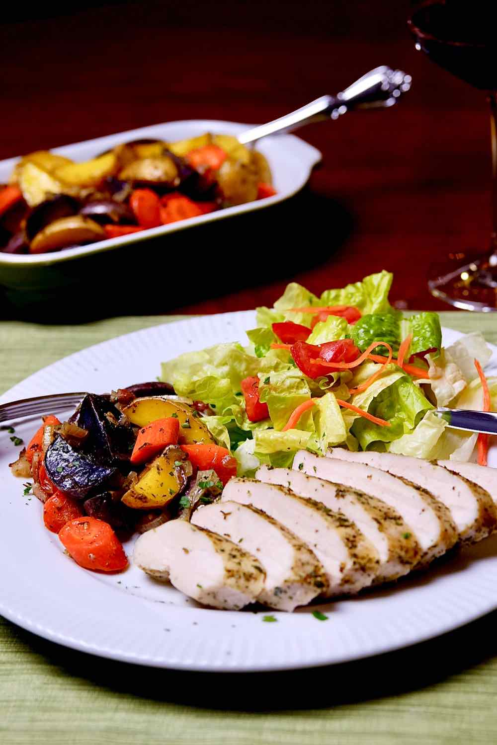 Lemon-Herb Roasted Chicken with Colorful Baby Potatoes and Carrots and salad on a white plate.  Wine glass and serving dish with additional potatoes in background.