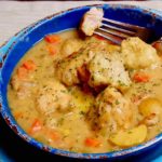 Light Chicken and Dumplings in dark blue bowl with bite shot of chicken on fork. Light blue charger in background set on a beige linen cloth.