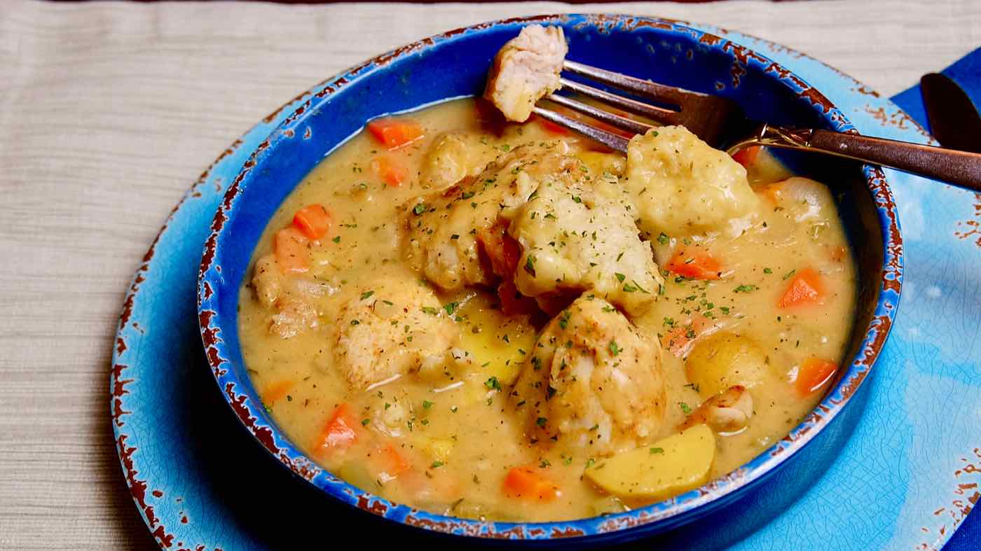 Light Chicken and Dumplings in dark blue bowl with bite shot of chicken on fork. Light blue charger in background set on a beige linen cloth.