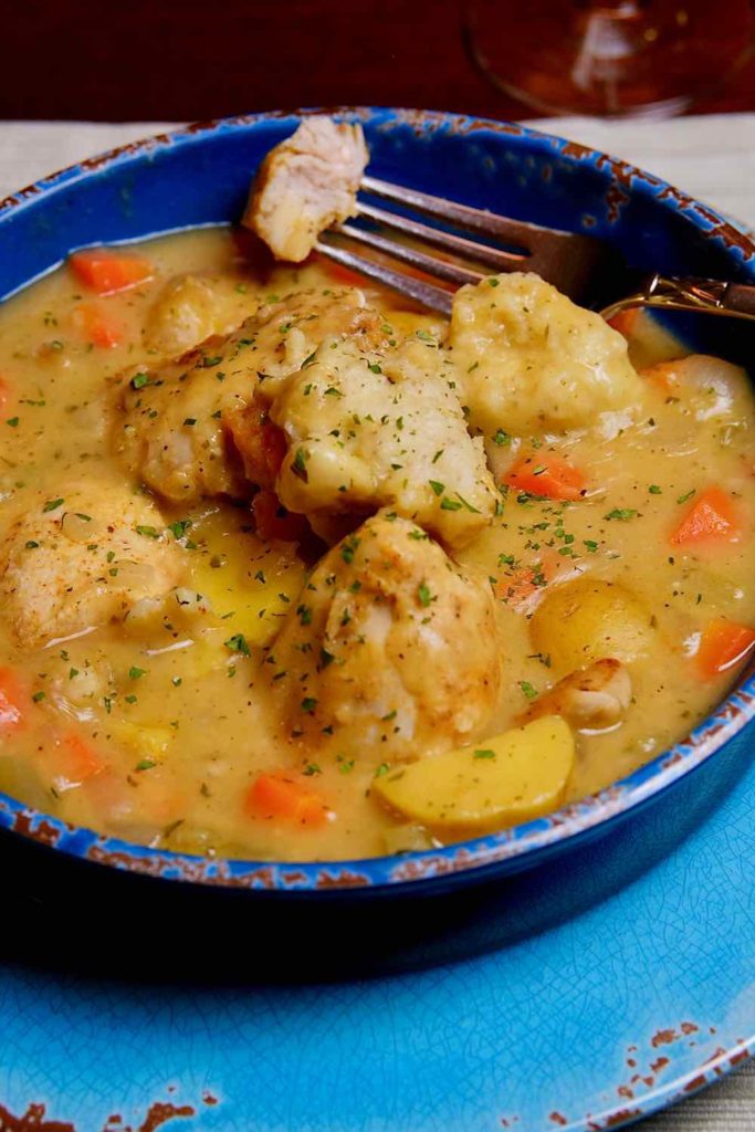 Light Chicken and Dumplings in a dark blue bowl with a fork bite of chicken, set on a light blue charger with beige linen placemat underlay.