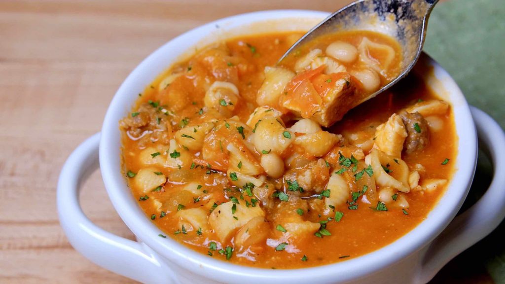 White bowl filled to brim with soup, spoon shot hovering over bowl with pieces of chicken, beans and pasta shells.