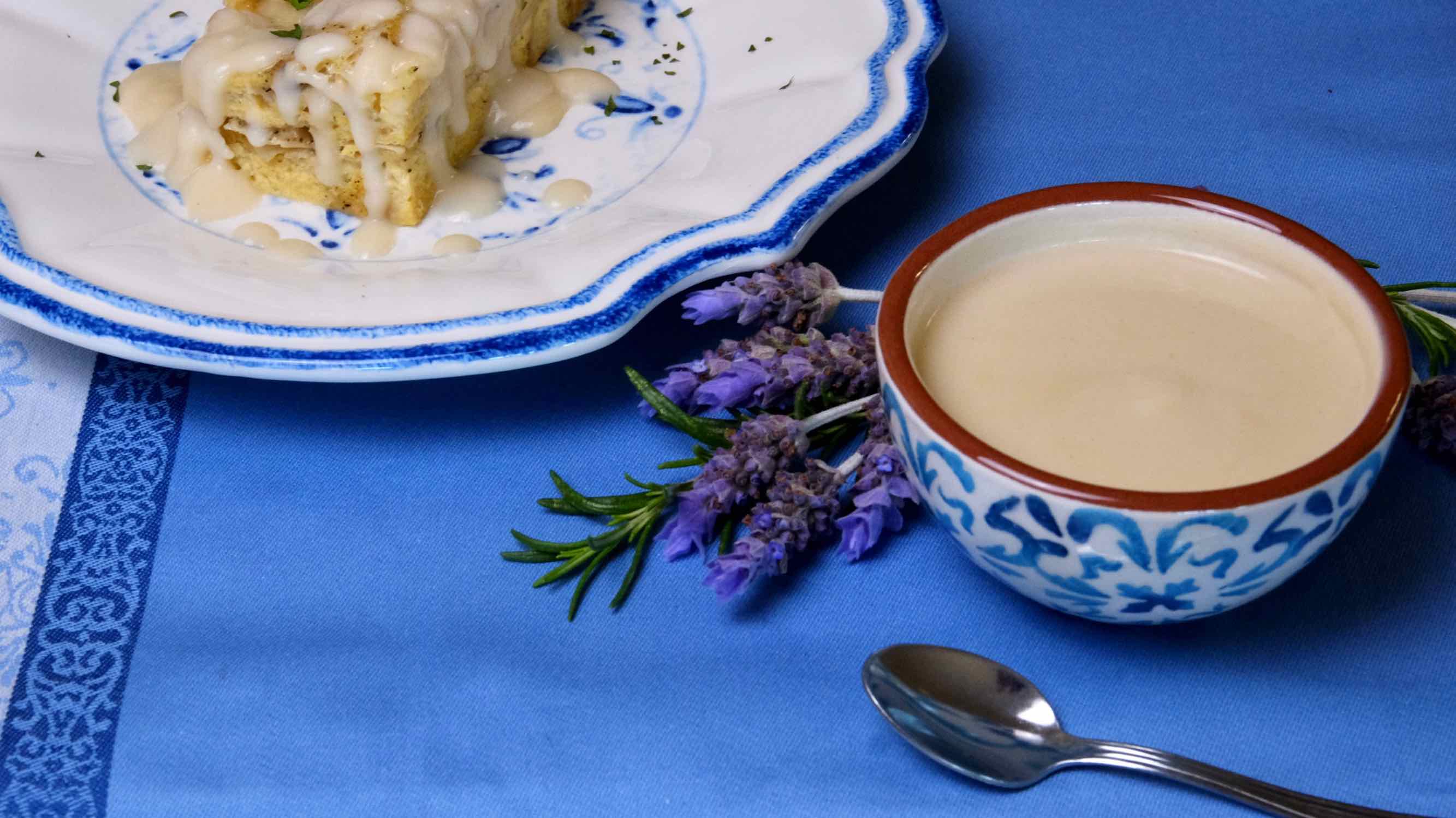 Light Creamy Parmesan Sauce with spoon in forefront. Lavender bouquet and dish with Ham and Gruyere Baked French Toast in background set on a periwinkle linen.