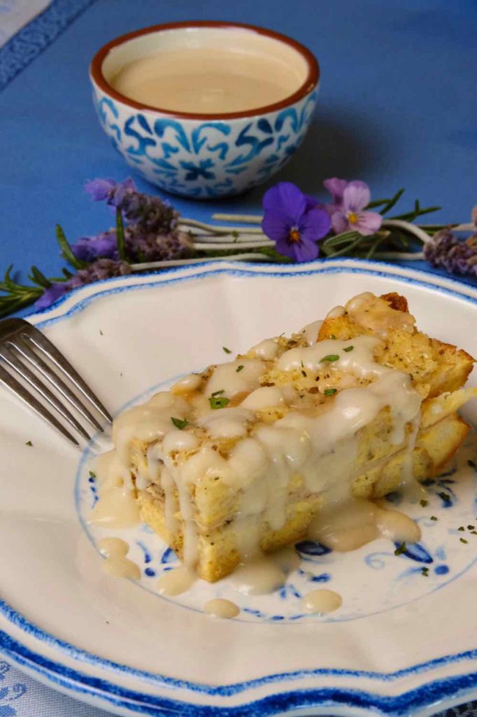 Light Creamy Parmesan Sauce drizzled over Ham and Gruyere Baked French Toast on a withe and blue plate with lavender bouquet and a blue and white bowl with additional sauce in background.