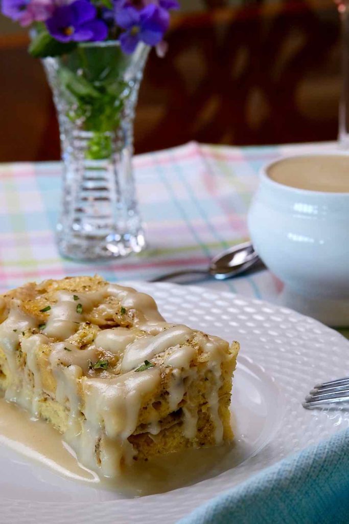 Light Creamy Parmesan Sauce drizzled over Ham and Gruyere Baked French Toast on a white plate with fork.  White bowl with additional sauce on a spring plaid linen underlay.