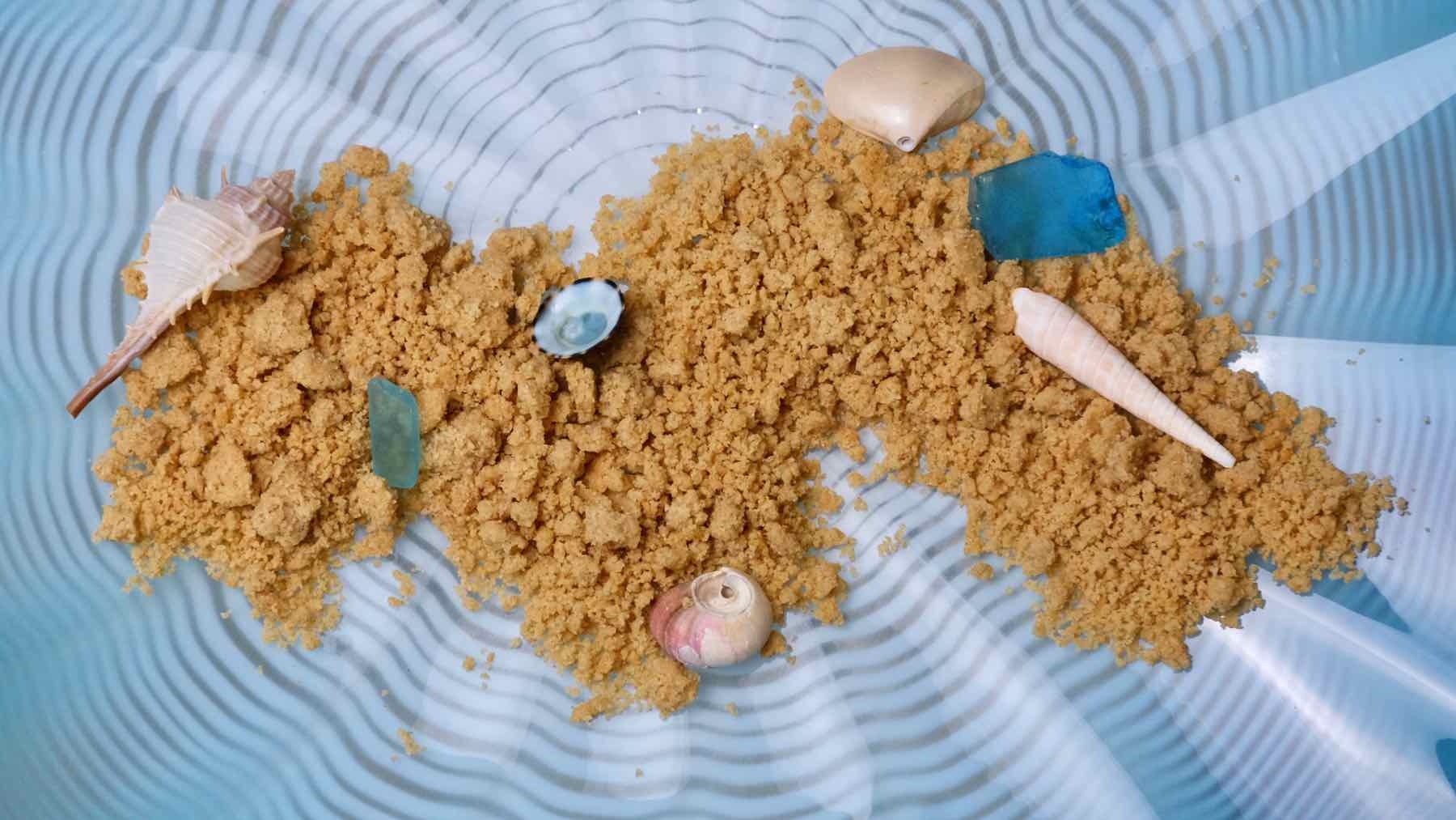 Easy Edible Sand scattered across a blue wavy dish and garnished with sea glass and shells.