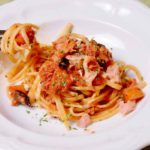 Easy Pasta Puttanesca with Tuna in white pasta bowl with a fork twirled with pasta in the left background.