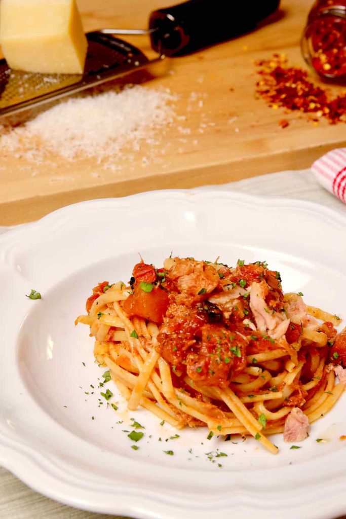 Easy Pasta Puttanesca with Tuna served in a white pasta bowl garnished with fresh parsley.  Cheese grater, grated cheese and chili pepper flakes on maple board in background.