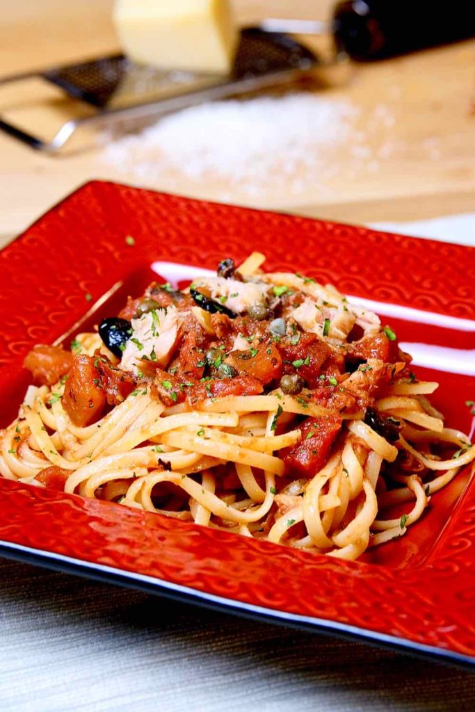 Easy Pasta Puttanesca with Tuna served on square red dish garnished with parsley.  Maple cutting board in background with cheese grater and a pile of grated Romano cheese.