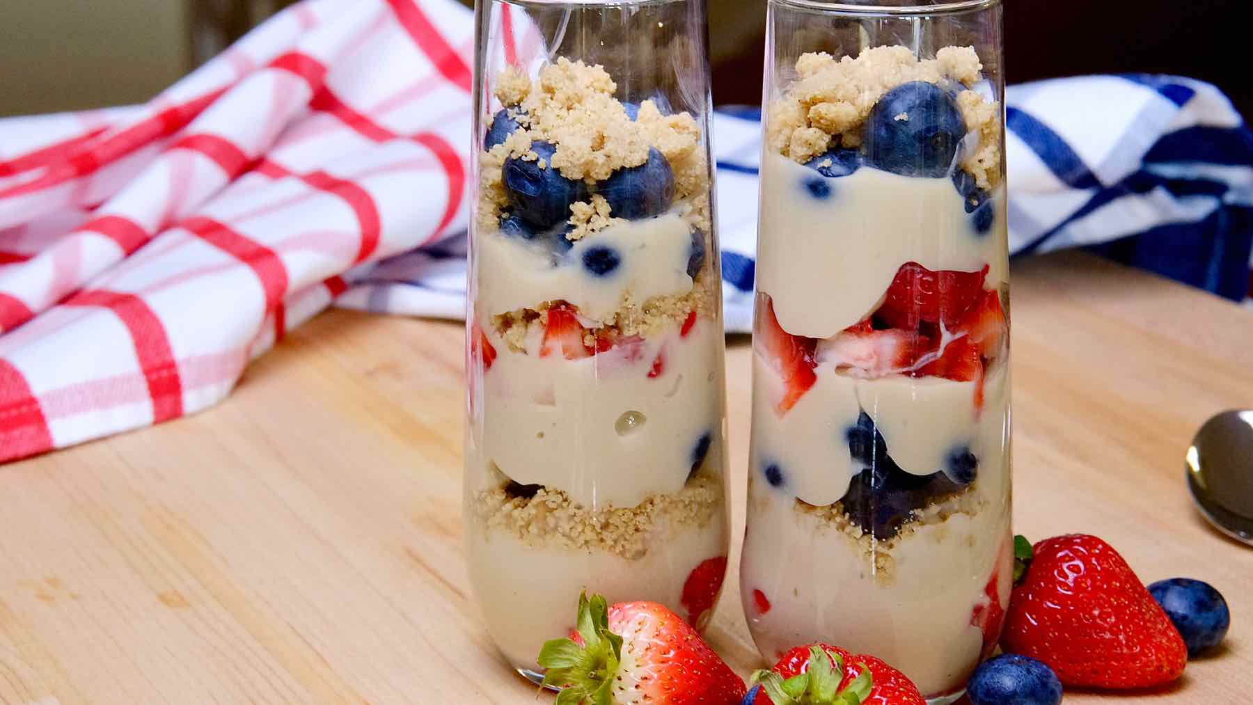 Red, White and Blue Berry Parfaits on maple board with fresh strawberries in foreground and red white and blue plaid linen in background.