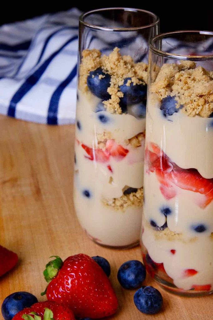 Red, white and blue berry parfaits layered in tall parfait glasses set on a maple board with fresh berries in forefront and red and blue plaid linen in background.