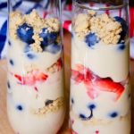Dairy free vanilla pudding with alternating layers of fresh blueberries, strawberries and edible sand in tall parfait glasses. Glasses are set on maple board with red and blue striped linens in background