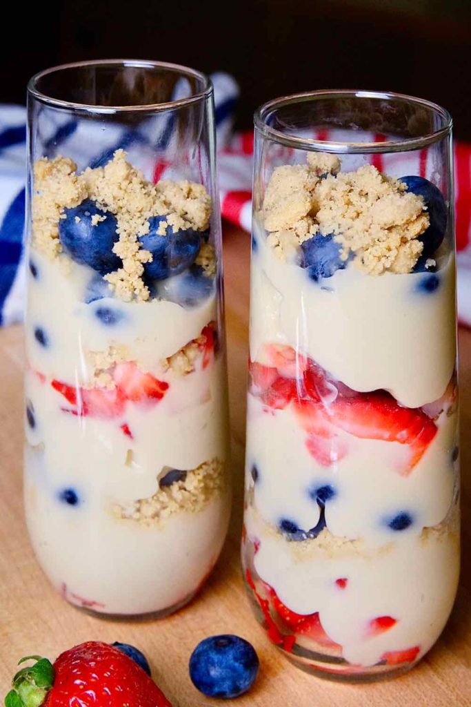 Dairy free vanilla pudding with alternating layers of fresh blueberries, strawberries and edible sand in tall parfait glasses.  Glasses are set on maple board with red and blue striped linens in background