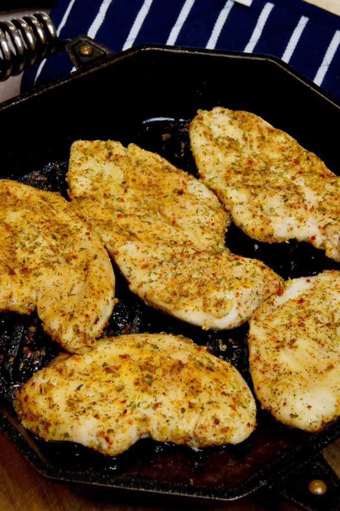 Easy baked chicken cutlets seasoned with Tuscan Herbs cooked in a cast iron griddle pan with navy and white pot holder in background.