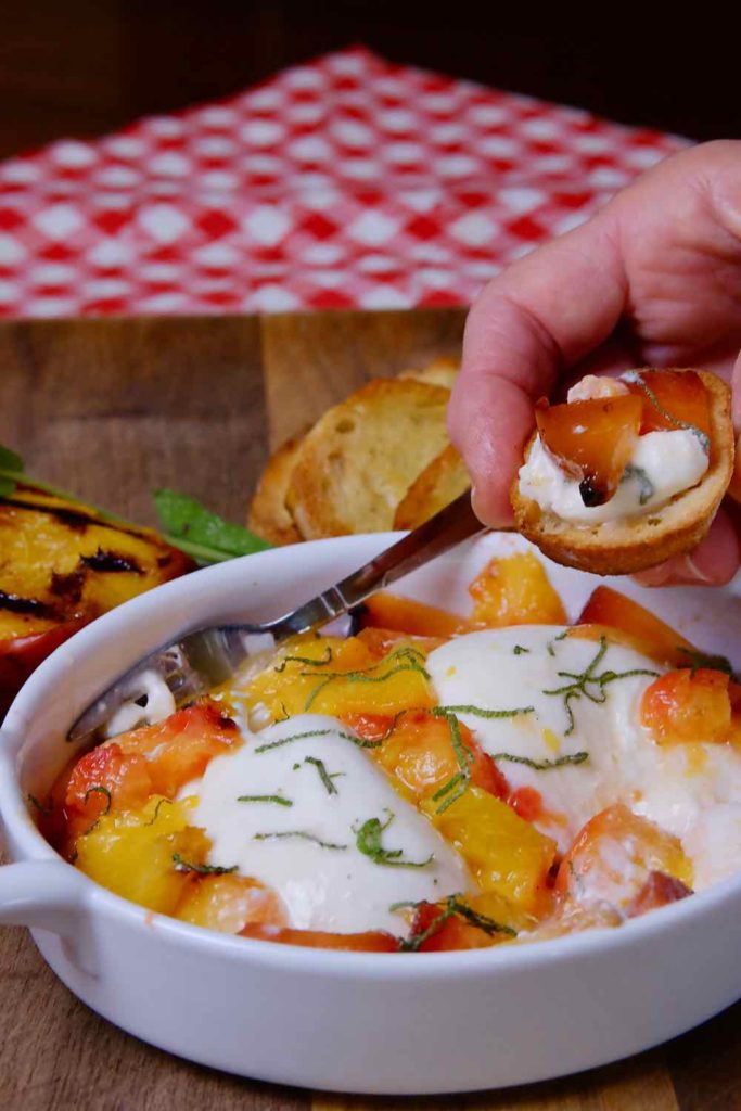 Burrata and Grilled Peach Appetizer in white serving dish set on walnut board with grilled peach and crostata in background.  A bite shot of crostata with burrata and peach is held in foreground.