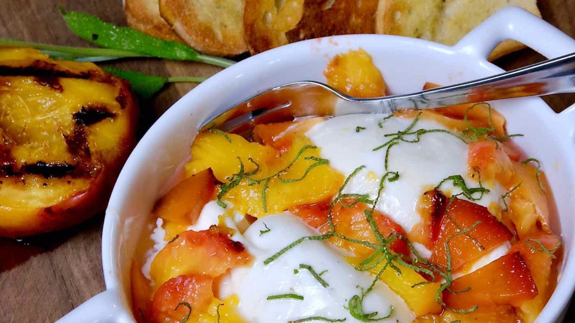 Burrata and Grilled Peach Appetizer in white serving dishing setting on walnut board with crostata and grilled peach in background.