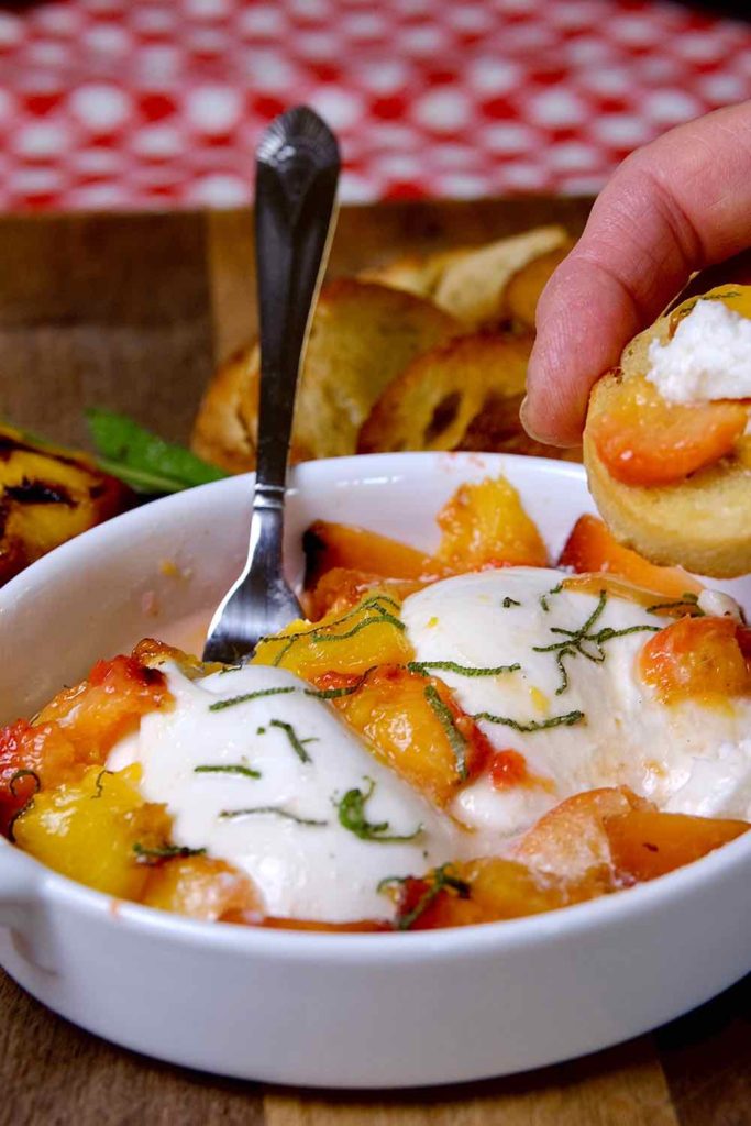 Grilled Peach and Burrata Appetizer served in a white oval terrine dish set on a wood board with red and white checked linen in background.