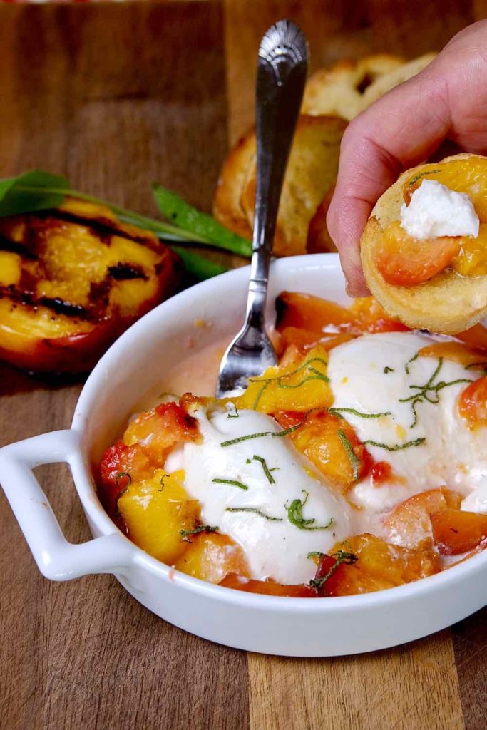 Burrata and Grilled Peach Appetizer in white serving dish with fork with a bite shot of crostata with peach and burrata being held. A grilled peach and crostata are setting in background on walnut board.