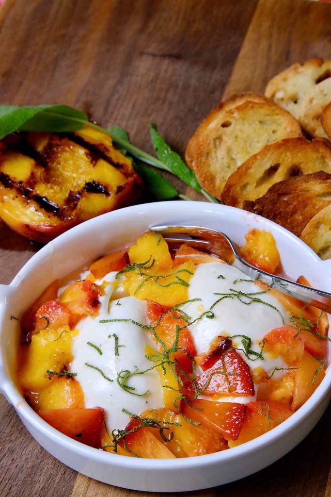 Burrata and Grilled Peach Appetizer in white serving dish set on walnut board with grilled peach and crostata in background.  