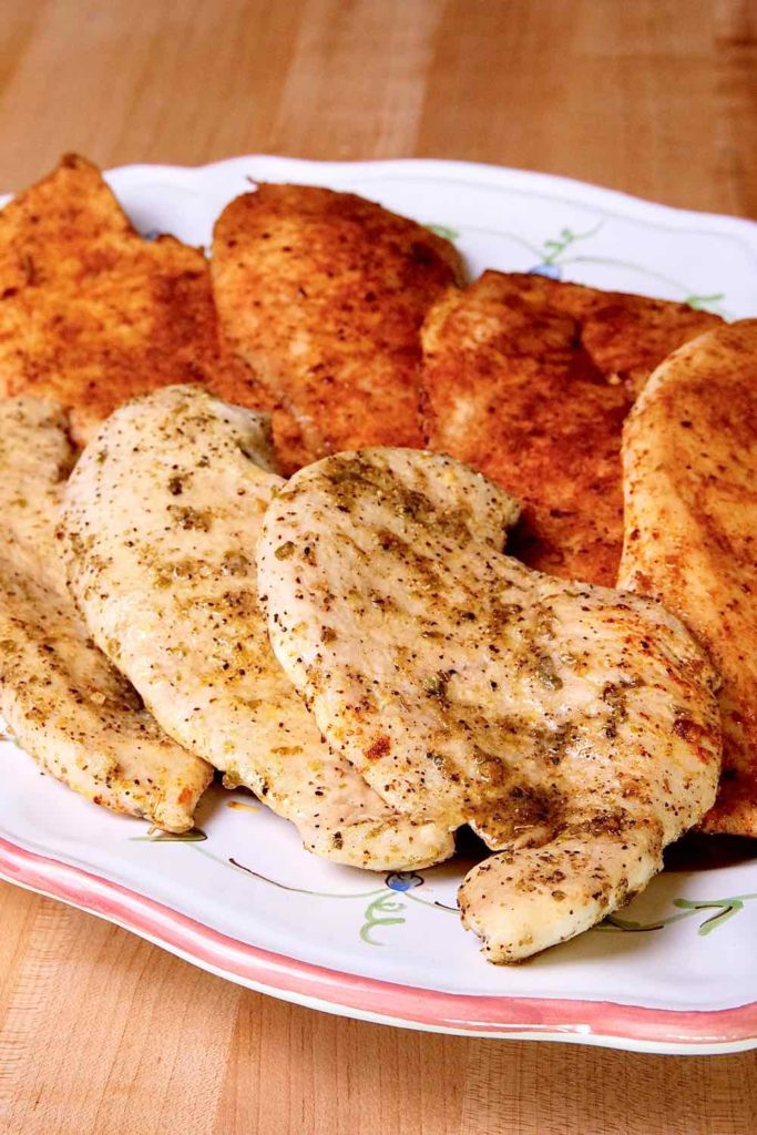 Easy Baked Chicken cutlets seasoned with Greek Seasoning and Barbecue Seasoning arranged on a white floral decorative platter setting on maple board.  