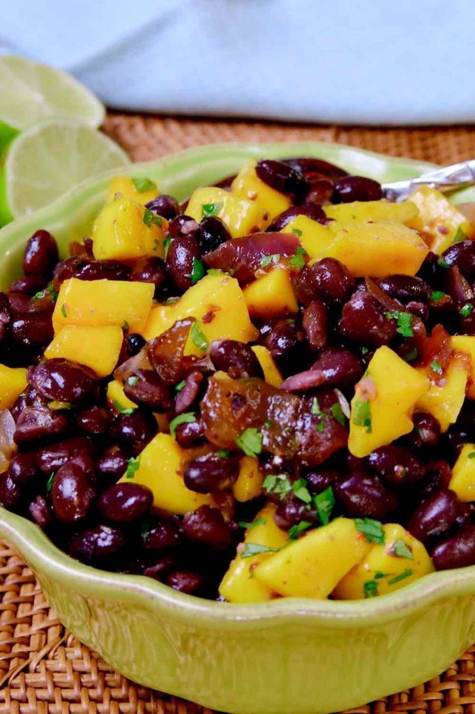 Black bean and mango salad served in a green bowl set on a rattan mat.  Blue linen and half of lime are sitting in background>