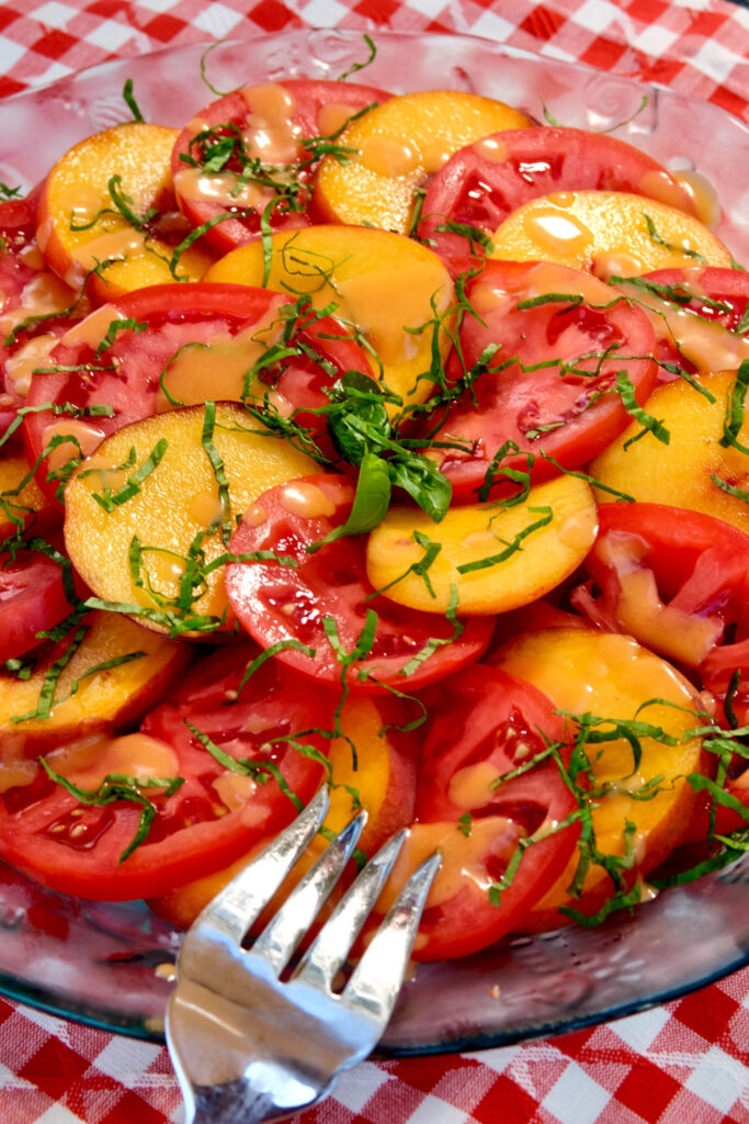 Slices of fresh peaches and tomato are layered in alternating slices around a serving platter, garnished with basil chiffonade and drizzled with fresh peach vinaigrette.  Platter is set on a red and white checkered linen with a serving fork perched on the edge of platter in forefront