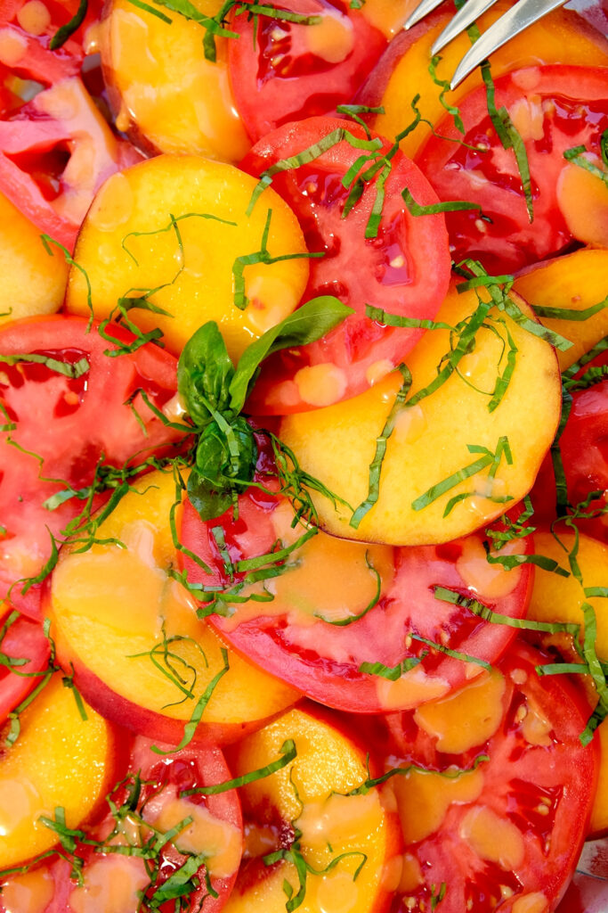 Overhead view of sliced fresh peaches and tomato are layered in alternating slices around a serving platter, garnished with basil chiffonade and drizzled with fresh peach vinaigrette.