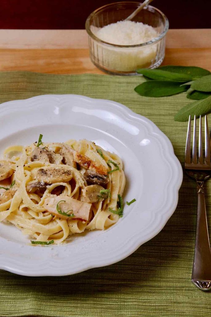 Light Chicken and Mushroom Fettuccine garnished with sage chiffonade in white bowl on green linen with utensils in foreground and bowl of cheese in background.