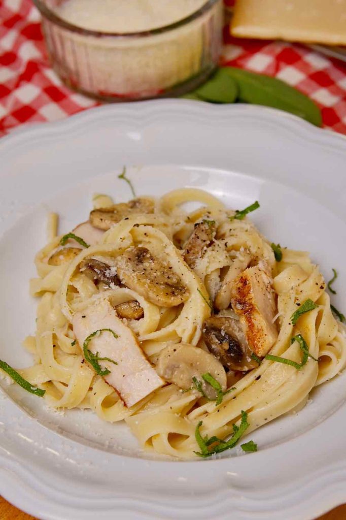 Chicken and Mushroom Fettuccine in white bowl garnished with sage.  Red and white checked linen and glass bowl of grated cheese in background