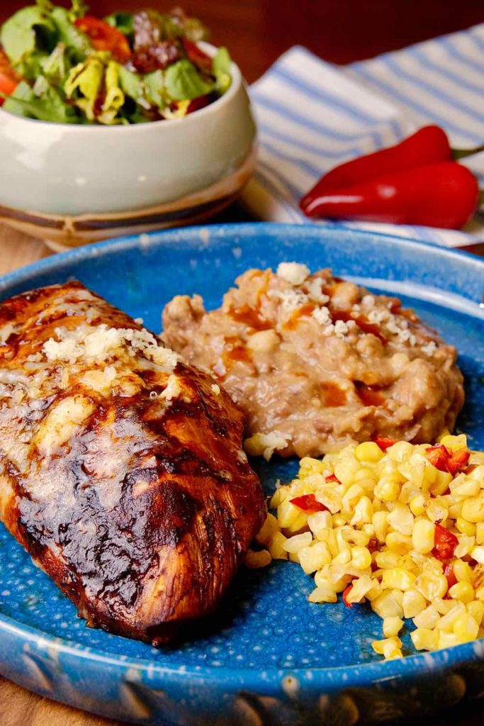 Guajillo Chili Chicken served on blue plate with Mexican corn and Skinny Refried Beans.  Side serving of salad and red chili peppers in background on blue and white linen.