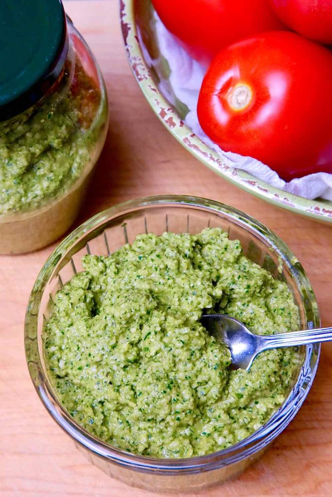 Jalalpeno Pesto Sauce in glass bowl with spoon set on maple board with jar of pesto and bowl of roma tomatoes in background.