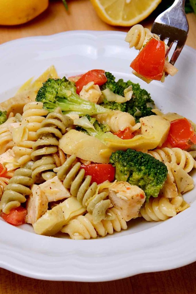 Pasta Salad Italian Style with broccoli, tomato, artichokes and cheese in a green bowl with lemons and parsley in background.