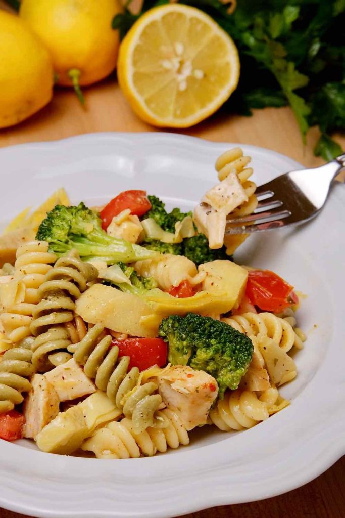 Pasta salad served in a white bowl with fork bite shot of pasta with chicken.  Lemons and parsley in background set on a wood cutting board.