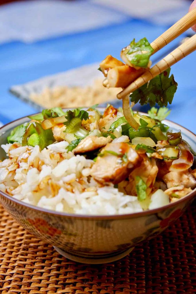 Healthy Hoisin Chicken Bowl with rice in floral blue rice bowl showing bite shot with chopsticks.  Small dish in background with chopped peanuts set on blue linen cloth.