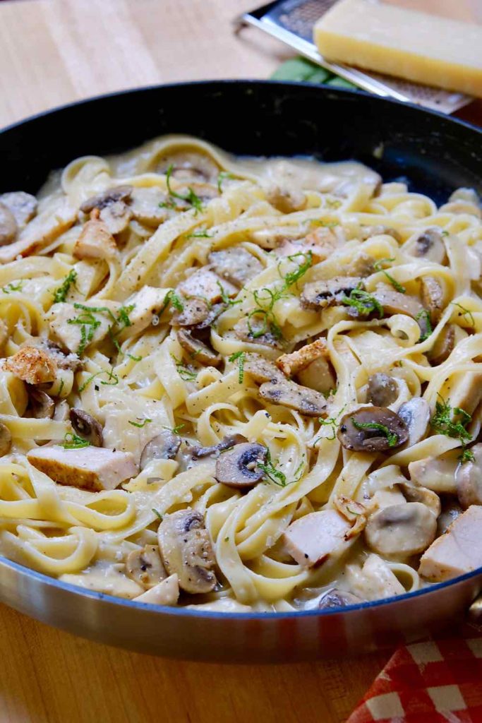 Light Chicken and Mushroom Fettuccine cooking in pan set on a maple board with red checked cloth in foreground and cheese grater in background.