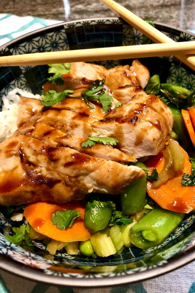 White rice, steamed carrots, snap peas and bok choy topped with sliced grilled Hoisin chicken is served in dark blue Japanese bowl.  Chopstick are crossed over top  rim of bowl 