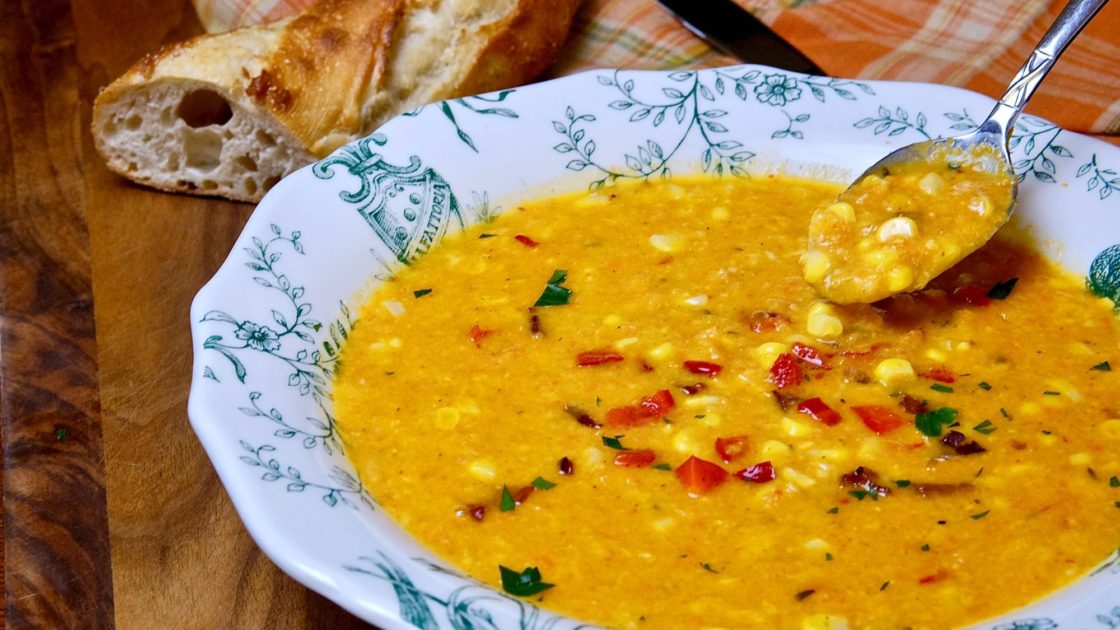 Hearty Creamy Corn Chowder with spoon full of chowder set over a white and green bowl set on walnut board with french baguette and orange plaid linen in background.