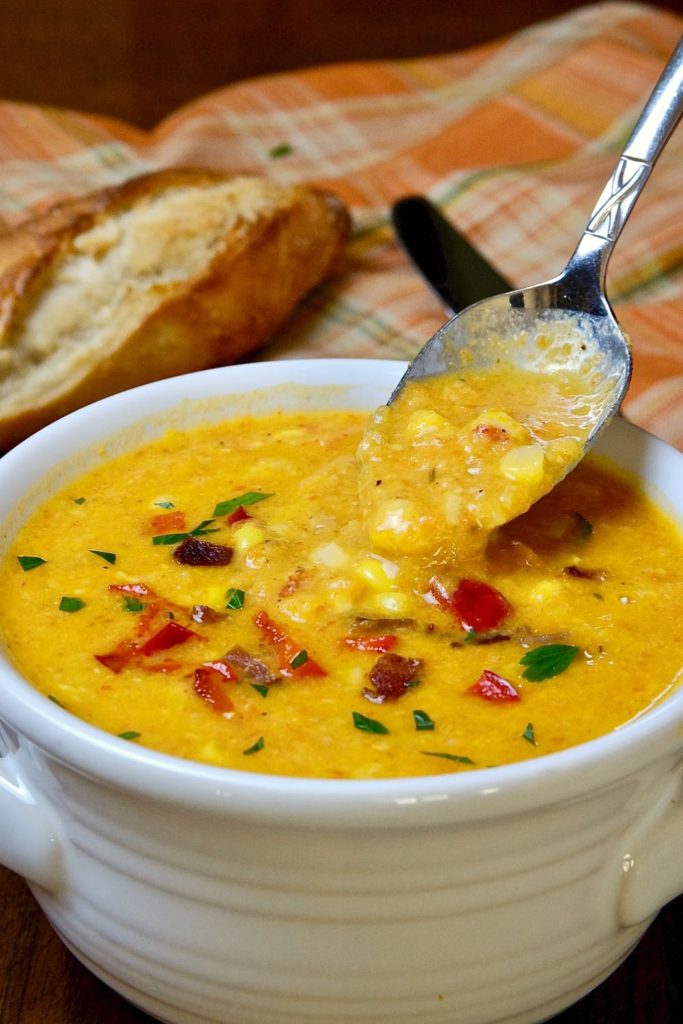 Hearty Creamy Corn Chowder in white soup bowl with spoonful of soup.  French baguette in bacground on a pumpkin colored linen.