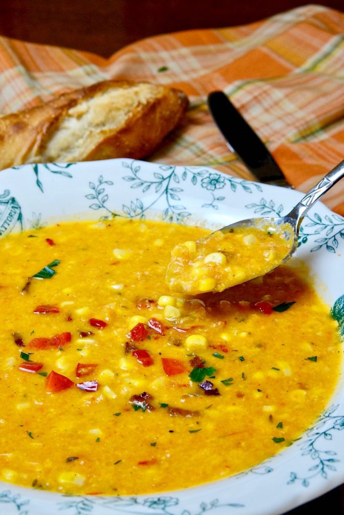 Hearty Creamy Corn Chowder in two-handled bowl featuring spoon shot of chowder. Chowder is garnished with diced red bell pepper, parsley and bacon bits. Orange plaid linen and French baguette in background.