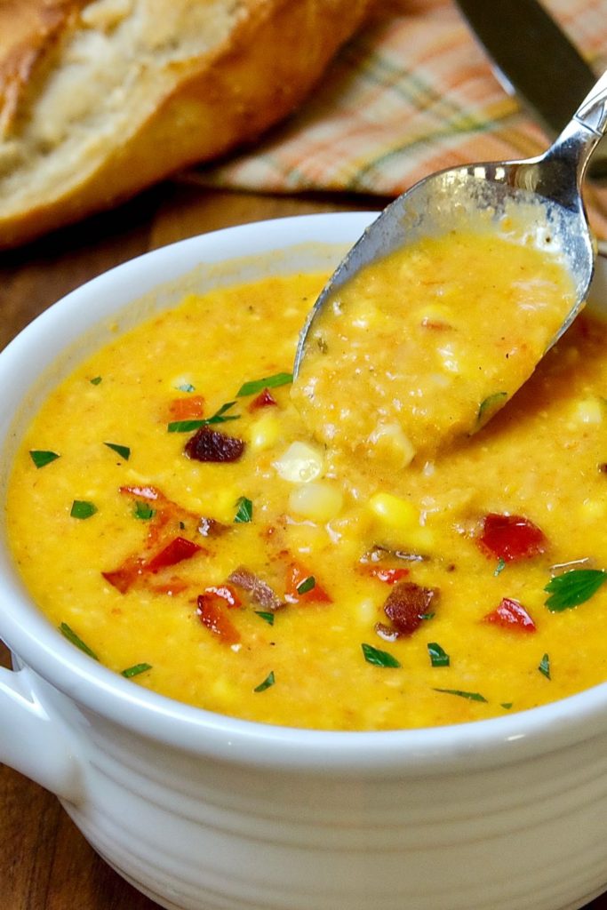 Hearty Creamy Corn Chowder in two-handled bowl featuring spoon shot of chowder.  Chowder is garnished with diced red bell pepper, parsley and bacon bits.  Orange plaid linen and French baguette in background. 