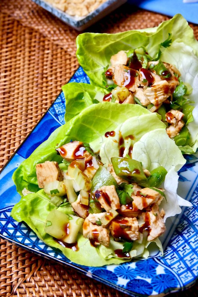 Hoisin Chicken and vegetables served on lettuce leaves set on blue patterned dish.  Rattan mat underlay with small dish of crushed peanuts in background.