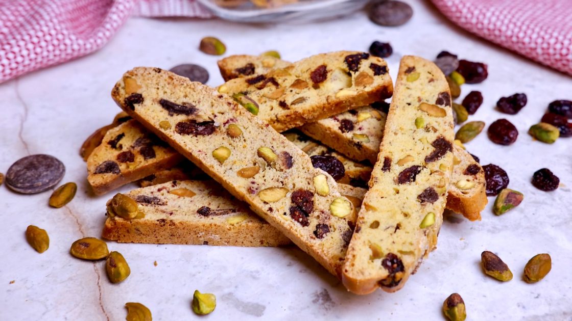 Chocolate Cranberry Holiday Biscotti on white marble with chocolate pieces, pistachios and cranberries scattered in foreground and red and white linen in background.