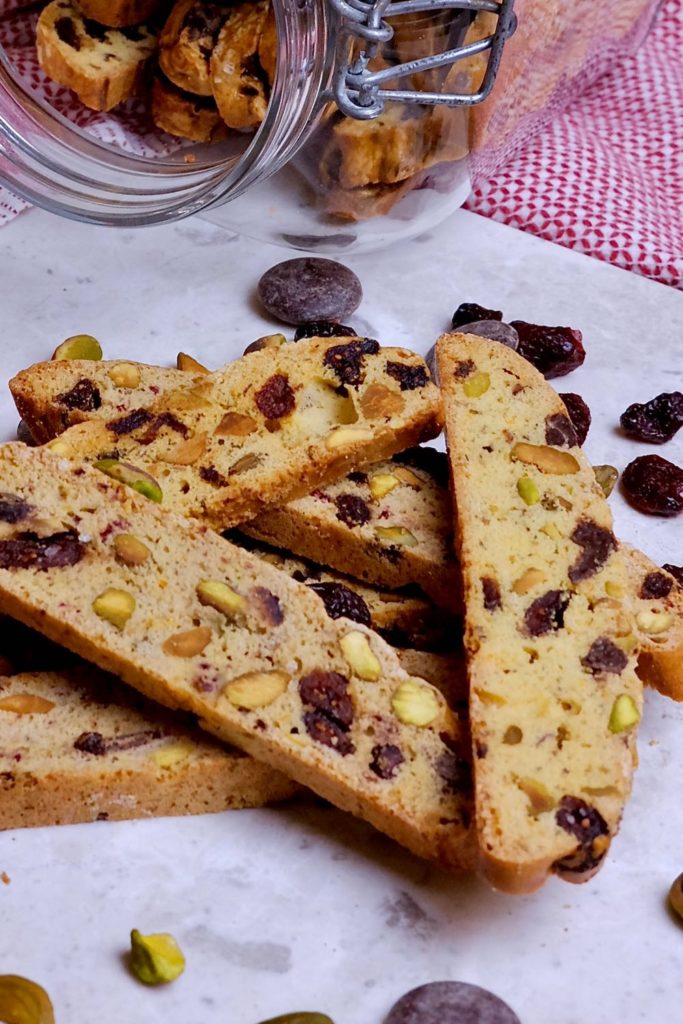 Chocolate Cranberry Holiday Biscotti displayed on white marble with cranberries, chocolate pieces and pistachios scattered around.  Red and white linen with glass cookie jar lying on side filled with biscotti.