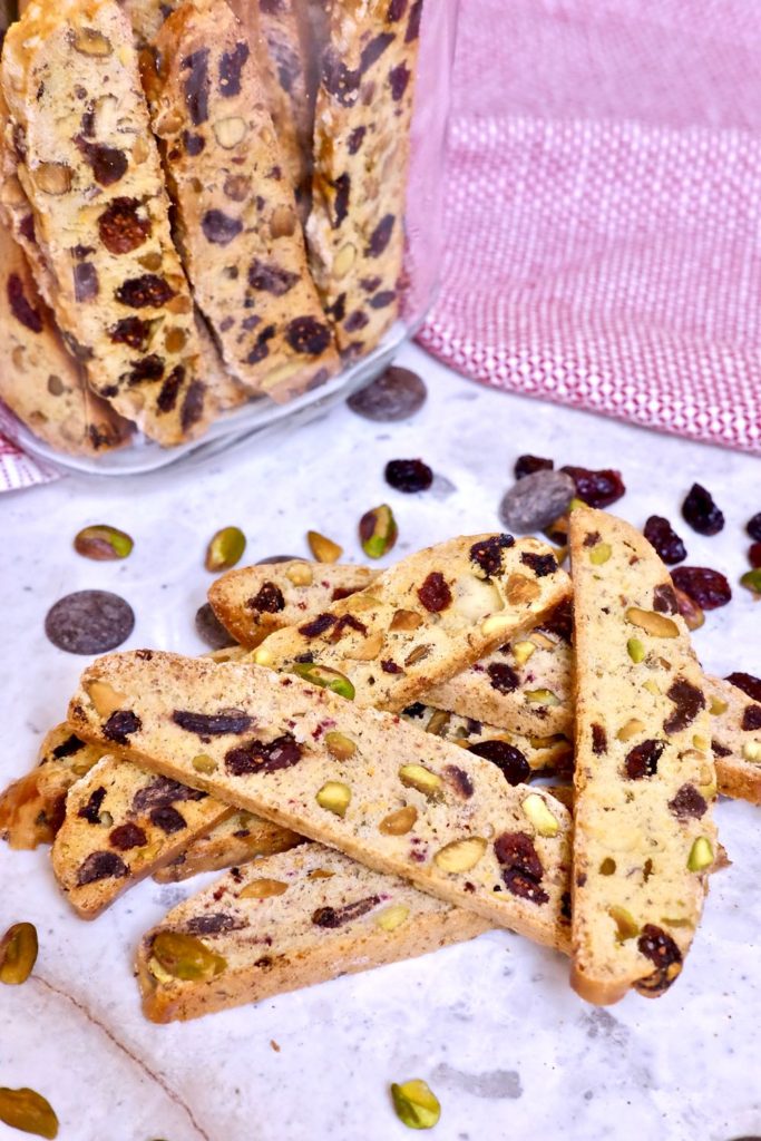 Chocolate Cranberry Holiday Biscotti displayed on white marble with chocolate and cranberries scattered around.  Glass jar of cookies and red linen in background.