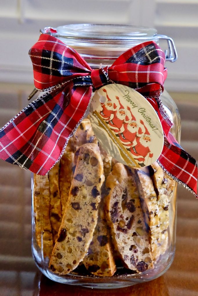 Chocolate Cranberry Holiday Biscotti in glass jar with red and black plaid bow and Christmas gift tag.  Jar is setting on a wood table.
