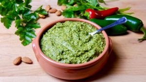 Jalapeno Pesto in terracotta serving bowl set on maple board with red and green chili peppers, cilanro and almonds scattered in background.