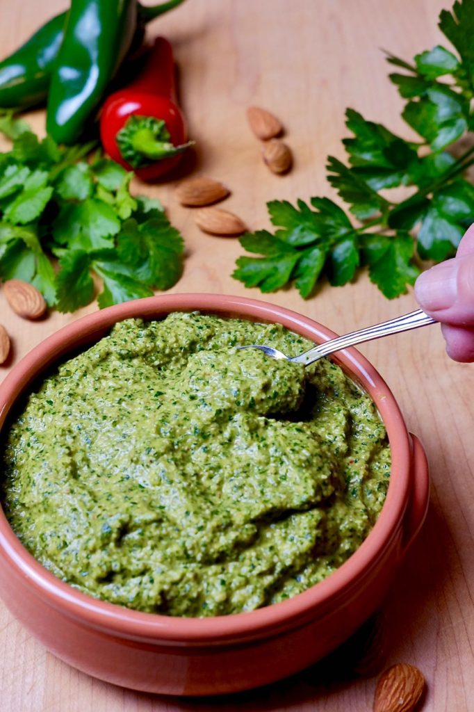 Terra cotta bowl of spicy pesto with fresh sprigs of cilantro, jalapenos and almonds in back ground.