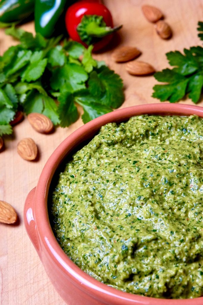 Spicy jalapeno pesto in terra cotta bowl set on maple board with fresh cilantro, parsley, chilies and almonds scattered in background.