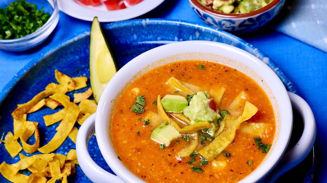 Chicken Tortilla Soup in white bowl set on blue plate with avocado and tortilla strips. Soup is garnished with tortilla strips, avocado and chopped cilantro.