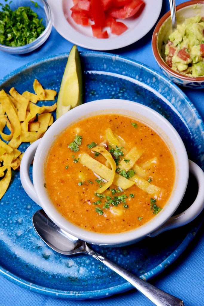 Creamy tortilla soup with chicken and garnished with tortilla strips. Condiment bowls of chopped avocado and tomato set in background.