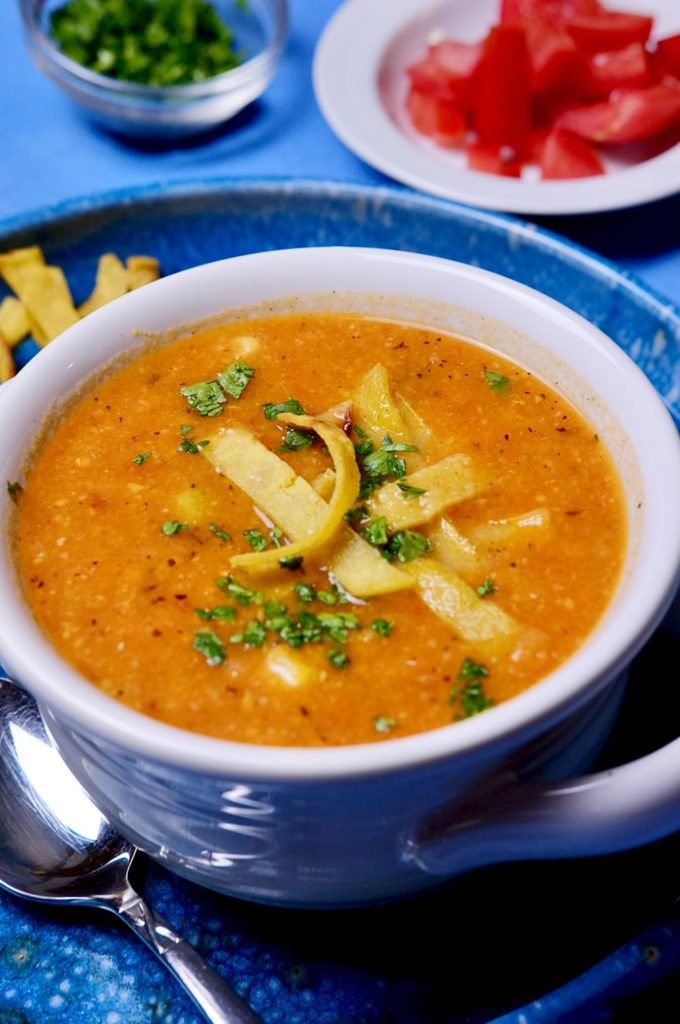 Creamy Chicken Tortilla Soup in white bowl set on blue plate garnished with tortilla strips.  Bowl of chopped tomatoes and cilantro in background set on blue linens.